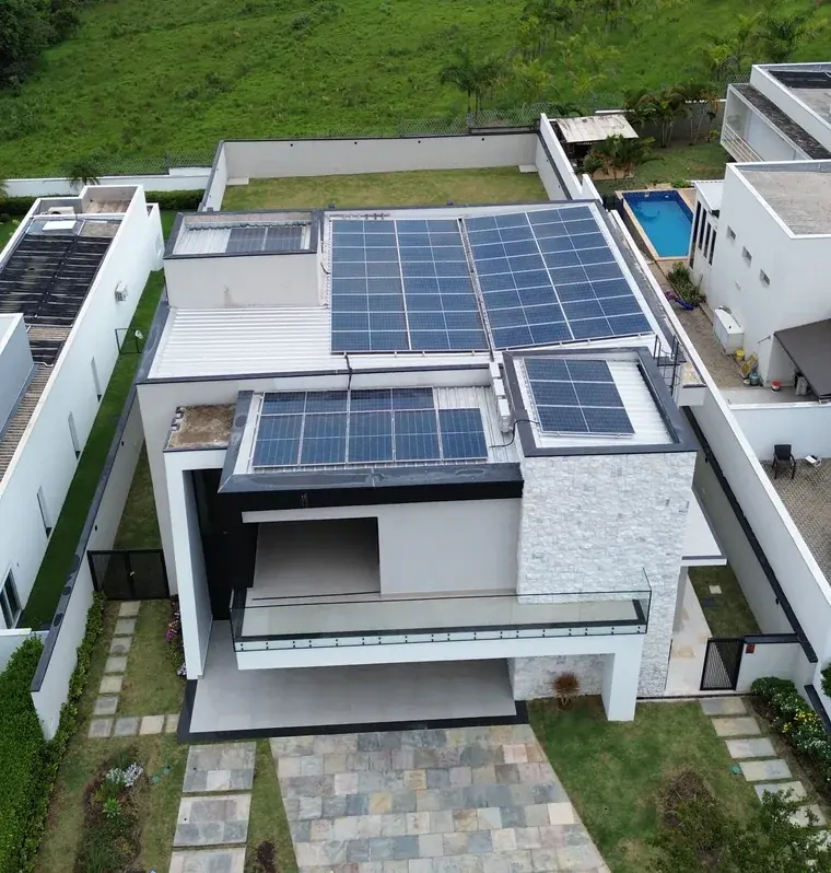 Vista aérea de drone de residência de alto padrão em condomínio de Sorocaba/SP, com 44 módulos fotovoltaicos espalhados por quatro telhados metálicos e campos verdes ao fundo